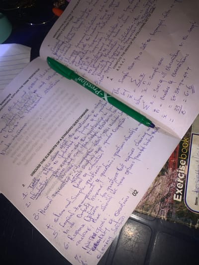 An overhead view captures an academic study setup located in Anantigha, Nigeria. The central subject is an open textbook or spiral-bound notebook displaying handwritten notes in blue ink across its two pages. A green "X-Pression" brand pen rests diagonally across the spine of the open book, with its tip pointing towards the upper left.
On the visible left page, the headings "EXERCISES" and "UNDERSTANDING EXPLAIN THE TERM 'STATISTICS'" are printed, accompanied by handwritten definitions related to statistics. The print on this page appears partially reversed due to the paper's transparency or underlying text.
The right page contains more extensive handwritten notes. At the top, a section titled "MENTION FIVE (5) TYPES OF FREQUENCY..." lists and briefly describes statistical concepts like "Grouped frequency distribution" and "Cumulative frequency." Below this, a problem asks to "COMPUTE THE MEAN for a Set of Ungrouped data" with given numbers (8, 5, 9, 4, 7). The solution demonstrates the mean calculation: "X = Sum of all Observations / Number of Observations = 33 / 5 = 6.6." Further down, a new section begins with "DISCUSS THE ELEMENTS OF A STANDARD QUESTIONNAIRE," followed by a numbered list including "1. Title," "2. Introduction," "3. Personal Information," "4. Clear and Simple Language," "5. Logical Arrangement," "6. Response Options," "7. Instructions," and "8. Preamble...Closing Section."
To the lower right of the primary book, a segment of another notebook, labeled "Exercisebook" with the partially visible name "Rosette..." and "Name:" field, is present. This notebook features a patterned cover with what appears to be a landscape or floral design. The background, particularly in the upper left corner, is dark and indistinct, showing hints of a light blue cup or container and some metallic objects. The scene is illuminated by an overhead light source, casting some shadows and highlighting the paper.