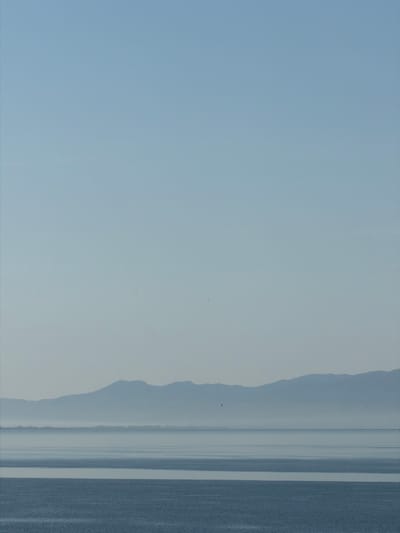 The image depicts a hazy, light blue-gray landscape. The right side of the frame features a dark gray strip that appears textured, suggesting land or a developed area. Adjacent to this is a lighter gray area, possibly water or a flat plain, with vertical lines hinting at reflections or distant structures. The left side of the frame is predominantly a light blue-gray sky. In the middle distance, there is a faint suggestion of land with undulating ridges or hills. There is no visible evidence in the photograph to pinpoint the location in Nador, Morocco, or of people, subjects, objects, actions, or interactions.