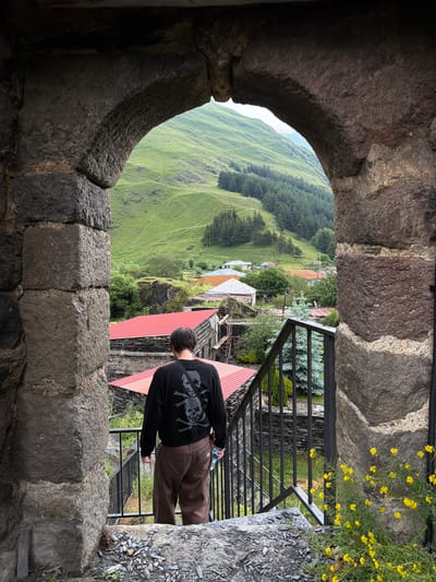 The image shows a view through a stone archway looking out onto a village nestled in a green, mountainous landscape. A person wearing a black sweatshirt with a skull design on the back and brown pants is standing on a stone staircase, facing away from the camera and looking out at the village. The village has red-roofed buildings and is surrounded by trees and rolling green hills. In the foreground, to the right of the staircase, there are yellow flowers. The stone archway is made of large, rough-hewn stones. The image captures a scenic view of a rural village, likely in a mountainous region like Sno, Georgia.