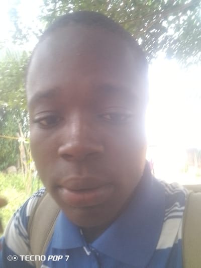 A close-up, outdoor, selfie-style photograph features a young person's face, which takes up most of the frame. The subject has dark skin and short, dark hair. They are looking directly at the camera with a neutral expression. The lighting is very bright, especially on the right side of the image, suggesting it is daytime and potentially sunny. The background is blurred and appears to be natural scenery, with green foliage and some branches visible, indicating an outdoor setting. A portion of the subject's blue and white striped shirt and a tan backpack strap are visible in the lower part of the image. The words "TECNO POP 7" are overlaid on the bottom left corner of the photograph, indicating the device used to take the picture. The location, Eleweran, Nigeria, suggests an urban or semi-urban setting with natural elements.