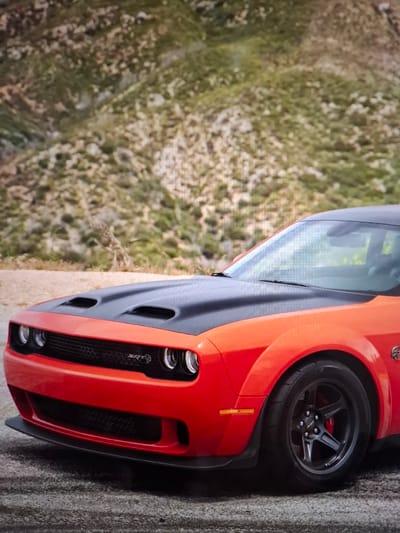 The image shows a close-up, low-angle view of the front portion of a bright orange Dodge Challenger SRT Hellcat. The car is parked on a paved surface, possibly a road or asphalt. The background consists of a dry, rocky hillside with sparse green vegetation. The car's hood is black and features prominent air intakes. The grille has "SRT" badging. The front bumper is also orange, with a black lower lip spoiler. The front wheel, visible on the right side of the frame, is black with a multi-spoke design. Red brake calipers are visible behind the wheel spokes. The windshield and part of the roof are also visible, reflecting the sky and surroundings. There are no people or other vehicles present in the image. The scene appears to be outdoors, in a somewhat arid or mountainous environment. There is no information in the image to suggest it is located in Argun, Russia.