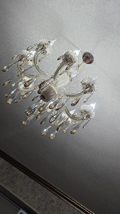 The image shows a close-up, low-angle view of a chandelier hanging from a textured, light gray ceiling. The chandelier is ornate, with multiple arms that curve upwards. Each arm terminates in a decorative holder with cut-out details and appears to hold a small, white lightbulb. Dangling from the arms are numerous faceted crystals, varying in size and shape, which catch the light. The chandelier itself has white and metallic elements, with some darker, intricate scrollwork on the holders. The ceiling has a subtle, almost fabric-like texture. Below the chandelier, a darker band runs horizontally, with a patterned wallpaper visible on the lower portion of the image. The lower right corner of the image is dark and out of focus, suggesting the presence of a wall or corner. There are no people or animals visible in the image. The lighting suggests the chandelier is illuminated, casting a soft glow. There is no information within the image to suggest a specific location within the city of Yandare, Russia.