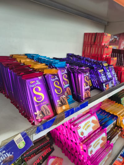 This image shows a close-up, eye-level view of a retail shelf stocked with an assortment of chocolates and wafers in a store located in Sidhpur, India. The primary focus is on rows of Cadbury Dairy Milk Silk chocolate bars, predominantly in purple packaging, arranged neatly on the shelf. Some of these bars are labeled as "Fruit & Nut," "Hazelnut," and "Oreo." Behind the Cadbury bars, stacks of red boxes, possibly containing more chocolates, are visible. To the right, there are also rows of other chocolate bars and wafer packets. On the lower shelf, partially visible, are more chocolates, including Toblerone bars, and colorful wafer packets in pink, yellow, and blue. The lighting suggests an indoor setting, likely a small grocery store or convenience shop. No people are visible in the frame.