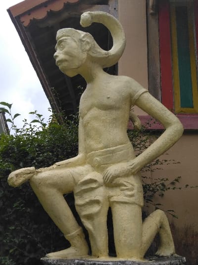 An outdoor, eye-level photograph captured in Ba Nwet Kone, Myanmar, depicts a pale yellow, granular-textured statue of a kneeling anthropomorphic figure positioned in the foreground. The figure, resembling a monkey-human hybrid, features a monkey-like face with large eyes, a snub nose, and a wide mouth. A thick, curved, snake-like appendage extends from the back of its head, arching upwards and backwards. The figure's torso is bare, revealing musculature, and it wears short, loose-fitting breeches gathered at the waist. Its left leg is bent with the foot flat and knee raised, while the right leg is bent with the knee on the ground and foot extended backward. The left hand rests on the left knee, and the right hand is placed on the lower back/hip.
Behind the statue, dense green foliage occupies the mid-ground on the left. To the right, a building wall, painted a light orange-beige, features a window or door frame with vertical panels in red, yellow, and a darker color. Above this, a brown roof overhang with scalloped eaves is visible. The sky in the upper left background is overcast with visible clouds. No people or direct interactions are visible.