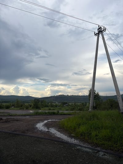 The image depicts a rural landscape near Skovorodino, Russia, under a cloudy sky. A prominent power line pole, constructed from two angled concrete pillars, stands on the right side of the frame, supporting high-tension wires. In the foreground, a dirt road curves, with a small stream of water flowing across it. Beyond the road, a grassy field extends towards a line of rolling hills covered in trees and vegetation. Small structures, possibly houses or outbuildings, are visible scattered in the distance amongst the greenery. The overall scene suggests a quiet, somewhat unpopulated area, likely on the outskirts of Skovorodino. There are no people or animals visible in the image.