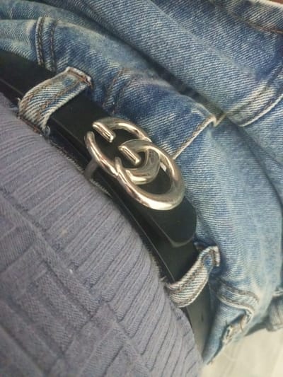 A black leather belt with a silver interlocked "G" buckle is worn with blue denim jeans. The belt and jeans are partially covered by a soft, light blue ribbed fabric, possibly a shirt or sweater. The scene is captured from a close-up perspective, focusing on the texture and details of the clothing and belt buckle. There is no discernible location in La Tagua, Venezuela.