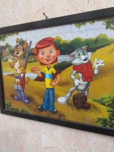 This is a framed jigsaw puzzle depicting a cartoon scene with three characters in an outdoor setting. On the left is a brown anthropomorphic dog wearing a green hat, a blue bandolier with bullets, a brown satchel, and large purple boots. The dog is holding a rifle and waving with its right hand. In the center is a cartoon child with red hair, wearing a yellow shirt with blue stripes and blue pants. The child is holding a circular, flat, reddish-brown object, possibly a cookie or pie, in their left hand. To the right is a grey anthropomorphic cat wearing a red scarf and brown boots. The cat is standing with its right hand on its hip and its left hand raised, also holding a small reddish object. The background of the puzzle shows a yellow-green grassy field with some trees and buildings in the distance, suggesting a rural or park-like environment. The puzzle is framed with a black border. The image is taken at an angle, showing the puzzle hanging on a wall with a textured, light-colored surface. There is no discernible location in the city of Samara, Ukraine.