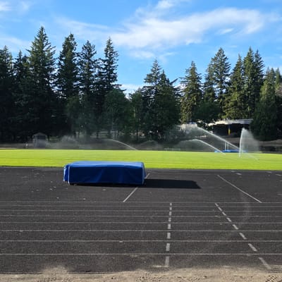 The image shows a blue tarp-covered object on a black running track. In the background, a green grass field is being watered by several sprinklers, with a line of evergreen trees behind it. A building with a light-colored facade and a dark roof is visible through the trees. The sky is blue with scattered white clouds. The location is the Roosevelt High School track and field in Portland, Oregon. There are no people visible in the scene.