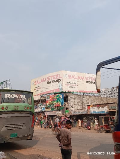 The image depicts a busy urban street scene in Kaliganj, Bangladesh, recorded at 14:06 on April 12, 2025, under a clear, light blue sky. In the foreground on the left, the rear of a green bus with "ROYAL EXPRESS" text and a "HINO" logo is visible, displaying a license plate ending in "0892". Debris is present on the ground next to the bus. On the right foreground, the partial front-right section of a blue and red bus is visible. Centrally in the mid-ground, a young man with dark hair, wearing a light brown t-shirt and dark pants, stands with his back partially to the camera, looking slightly to his left, holding an object in his left hand behind him. The road surface is unpaved and dusty. Multiple pedestrians are present, walking and standing, alongside several rickshaws and auto-rickshaws, including a red auto-rickshaw on the far right. In the background, a multi-story commercial building features prominent red and white signage for "SALAM STEEL THERMEX" and Bengali text, including "উন্নত এনফোর্মা" (Advanced Enforma). Below this primary signage, numerous colorful billboards and shopfronts line the street, displaying various advertisements, including an "OPPO" logo.