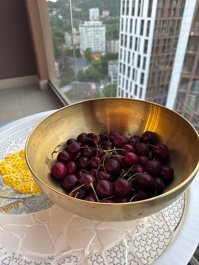 A close-up shot shows a golden bowl filled with dark red cherries, some of which have small water droplets on them, placed on a decorative tiled surface. To the left of the bowl, a portion of a yellow mosaic pattern is visible. In the background, through a window, a city skyline can be seen with various buildings, including modern high-rises and older structures, surrounded by greenery. The scene appears to be set on a balcony or a similar outdoor space, offering a view of the city of Vardane Microdistrict, Russia, possibly during the evening due to the lighting. There are no people visible in the image.