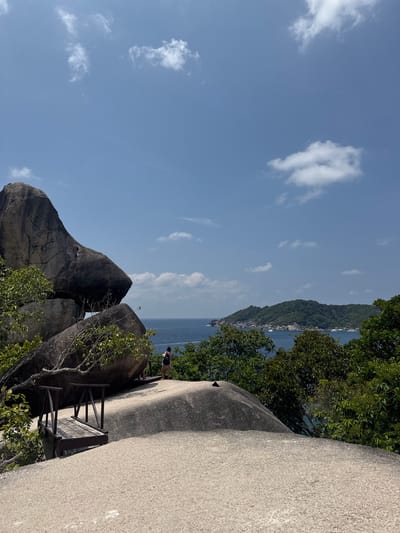 A photograph depicts a scenic overlook in Lam Kaen, Thailand. It features large granite boulders in the foreground, partially covered with greenery. A small wooden observation platform is affixed to one of the boulders. A person stands on the edge of the boulder looking out towards the sea and an island in the distance. The sky is blue with scattered clouds. The photograph appears to be taken from a low angle, emphasizing the height of the rocks.