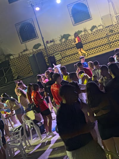 The image depicts an outdoor gathering at night, with a crowd of people of various ages in what appears to be a street or plaza. There are buildings in the background with windows covered by decorative latticework. Stage lighting, with a prominent blue hue, illuminates the scene. Some individuals are dancing or socializing. A woman in a red top and black skirt is visible on an elevated platform or balcony in the background. Empty plastic chairs are in the foreground, suggesting a seating area that is currently unoccupied or has been vacated. The overall atmosphere suggests a lively event, possibly a festival or party. The location is identified as Blanca, Spain.