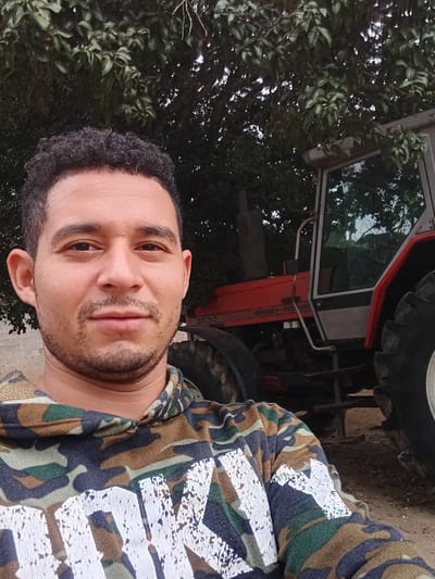 A selfie of a man with short dark curly hair and a trimmed beard wearing a camouflage shirt with white geometric designs. He is standing in front of a red Massey Ferguson tractor, partly obscured by foliage. The scene appears to be outdoors, potentially on a farm or rural area. The specific location in Cujisal, Venezuela cannot be determined from the image.