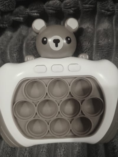 The image shows a bear-shaped "pop it" fidget toy resting on a textured surface, possibly a blanket or fabric. The toy is primarily light in color, with a bear head featuring eyes, a nose, and ears. Adjacent to the bear's head is a rectangular section containing a grid of circular "pop it" bubbles. The toy has several buttons on its side. The scene is in black and white. The location, Villa Ballester Noroeste, Argentina, is not visible in the image.