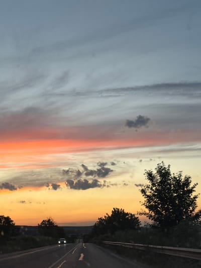 This media file captures a picturesque sunset scene, likely taken from a moving vehicle on a road. The sky dominates the frame with a gradient of colors, transitioning from a soft blue-grey on the left to warm oranges and pinks on the right, indicative of twilight. Wispy clouds are scattered across the sky, adding texture to the scene. Silhouetted trees line the right side of the road, their dark forms contrasting with the vibrant sky.
The road itself is visible, paved and marked with white lines. In the distance, a single car with its headlights on is driving away from the viewer. The overall mood is serene and tranquil, evoking the beauty of a natural sunset. There are no visible people or text in the image. The presence of the road and trees suggests a rural or suburban setting. The time of day is clearly sunset or dusk.