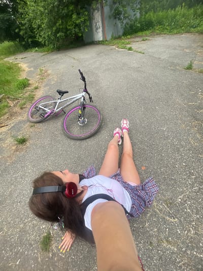 A young Caucasian woman with long brown hair, wearing red headphones, a white t-shirt, and a plaid skirt, lies on asphalt. A bicycle with pink rims is next to her. The scene appears to be outdoors, possibly in a park or abandoned lot, with trees and a metal building in the background. The location is specified as Frontoviki, Russia.
