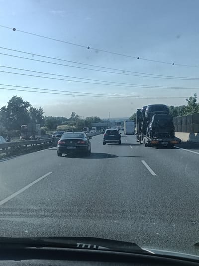 The image depicts a highway traffic scene on a bright, sunny day. In the foreground, a vehicle's windshield wiper is visible. The highway stretches into the distance, with multiple lanes of traffic. Several cars and trucks are present, including a car carrier truck loaded with dark-colored vehicles. In the background, trees and some structures line the side of the highway. The sky is clear with a few power lines visible overhead. The location is identified as the highway near Všechromy, Czechia. There are no visible people in the image.