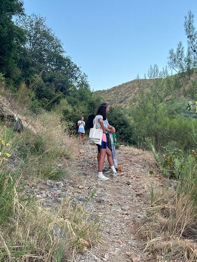 In Seoce, Montenegro, three young females are captured on a rocky, uphill path surrounded by dense greenery. The first female, in the background, wears a light-colored, sleeveless dress and stands facing left. Two other females are closer to the foreground, both looking to the right. The one on the left has dark hair and is wearing a light-colored t-shirt with a tote bag slung over her shoulder, dark shorts with a pink trim, and white sneakers. The female to her right wears a light blue t-shirt and dark shorts. The path is uneven and composed of stones and dirt, with dry grasses and green vegetation growing alongside it. The background features a steep, tree-covered hill under a clear, light blue sky.