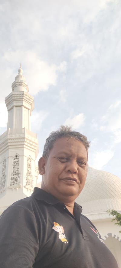 The image is a selfie taken outdoors in front of a mosque. A middle-aged man with graying hair and dark skin is positioned in the foreground, facing the camera with a slight smile. He is wearing a black polo shirt with a colorful cartoon graphic of an elephant on the left side of the chest. In the background, to the left of the man, is a tall, white minaret with intricate geometric patterns. To the right of the man is the white dome of the mosque. The sky above is partly cloudy and bright. The location is identified as Ranto, Indonesia.