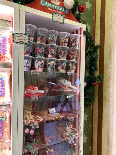 This media file presents an indoor view of a vibrant candy display, likely in a sweet shop or market, adorned with festive decorations. Above the main display, a green garland with small colorful lights and red spherical ornaments creates a celebratory atmosphere, possibly indicating the Christmas season. The background wall features a playful design with green stars and red and white stripes.
On the left side of the upper display, a red sign reads "CANDYTALE" and "Cirque du Bubble," accompanied by a price of "2.50€," suggesting a brand or product name and its cost. Below this, a white decorative item is visible. Further to the right, a clear sign in Greek states "ΑΠΑΓΟΡΕΥΕΤΑΙ Η ΔΟΚΙΜΗ," which translates to "No Sampling Allowed" or "Tasting Prohibited," reinforcing the commercial setting in Chalkida, Greece.
The main display features a wide array of confections. To the left, several rows of clear plastic cups are neatly stacked, each containing pre-packaged items labeled "Bubble Tale." To the right, multiple transparent acrylic dispensers are filled with various bulk candies. These include colorful, irregular-shaped gummies, red and white/yellowish candies (possibly gummy worms or slices), and assorted ring-shaped gummy candies, along with other sweets. White plastic scoops are placed inside the bulk candy bins for serving. Below this, the lower shelves show more pre-packaged sweets in clear bags. No people are visible in the image.