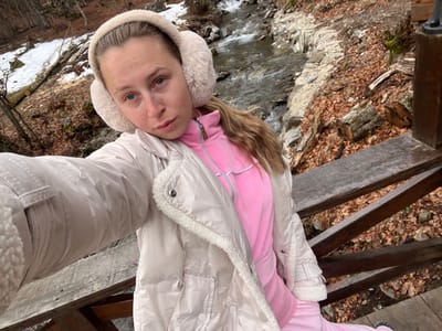 A female subject is photographed in a close-up, appearing to take a selfie. She wears a cream-colored puffer jacket with a sherpa-lined collar and cuffs, a pink full-zip tracksuit top, and matching pink tracksuit bottoms. Cream-colored furry earmuffs cover her ears, and her light-colored hair is partially visible over her right shoulder. Her left arm is extended towards the camera's perspective. She stands on a weathered wooden structure, with a railing visible in the foreground, below her. A flowing river with rocky banks is visible behind her, primarily to her right and in the background. The banks are covered with fallen brown leaves. Patches of snow are visible on the left bank and higher ground among deciduous trees, which appear bare. The scene is situated in Crkvine, Montenegro.