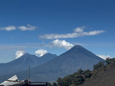 Three verdant mountains dominate the landscape under a bright blue sky dotted with fluffy white clouds. The largest mountain, situated to the right, features distinct vertical lines etched into its slopes. A smaller mountain is to the left of the large one, and one sits slightly behind it. In the foreground, hints of a roof and vegetation suggest a man-made structure or natural setting, grounding the majestic scenery with a touch of the familiar.