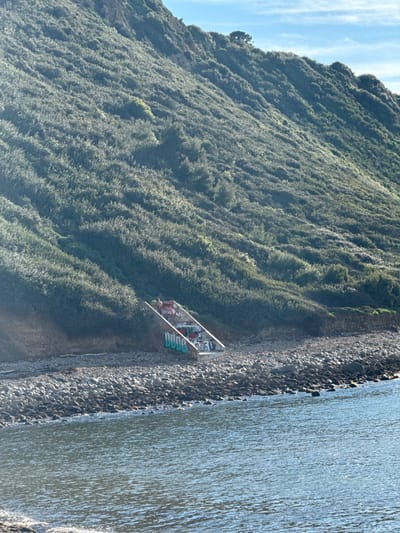 This image depicts a coastal scene on the coastline of Redondo Beach, United States of America, under clear daytime conditions. A steep, heavily vegetated bluff dominates the upper and mid-left frame, sloping downwards towards a rocky beach composed of dark stones and pebbles. Embedded into the base of the bluff, at its transition to the rocky shore, is a concrete staircase structure. This staircase descends towards the water and is extensively covered with multicolored graffiti, including the word "DUDE" prominently displayed in light blue letters on the lower visible steps. Two individuals are observed stationary at the top of the concrete staircase. The immediate foreground is calm ocean water, exhibiting ripples and reflecting ambient light. No discernible interactions between the individuals or with the environment beyond their stationary presence are evident.