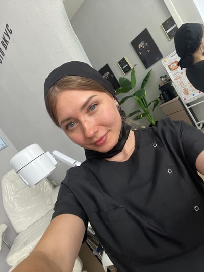 A young woman with blonde hair, blue eyes, and freckles is taking a selfie in a room that appears to be a beauty salon or medical office. She is wearing a black scrub top and a black head covering. A black face mask is hanging around her neck. She is smiling at the camera. In the background, there is a white chair covered in plastic, a white lamp, and various framed pictures on the wall. There is also a large plant and a whiteboard with anatomical diagrams. The room is clean and well-lit. The lettering on the wall to the left is in Cyrillic script. Based on the provided information, the location is identified as Stepnogorsk City Administration, Kazakhstan.