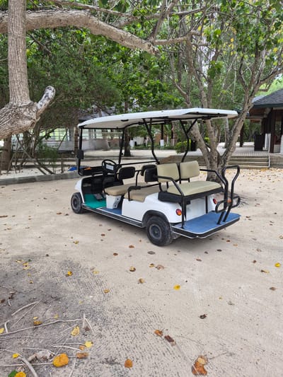 A white golf cart with beige seats sits parked on a sandy area. The cart is oriented towards the left of the frame. Behind it, a building with wooden stairs and a covered porch is partially visible. A tree with dense green foliage and exposed branches is also on the left, partially obscuring the building. The ground is sandy with scattered leaves. The scene is located in Huraa, Maldives.