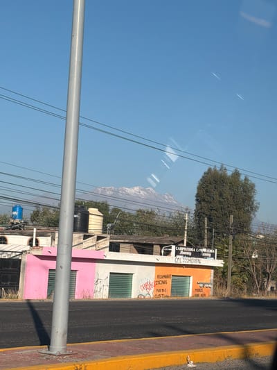 The image depicts a daytime street scene in Santa María Zacatepec, Mexico, with a prominent snow-capped mountain visible in the distance under a clear blue sky. A tall, light gray utility pole stands in the center-left foreground, partially obscuring the view. Behind this pole, a dark asphalt road occupies the mid-ground, bordered by a yellow curb and a reddish-brown sidewalk.
Across the street, two single-story buildings are visible. The building on the left features a pink lower facade with a white upper section and two closed green roll-down security shutters. Graffiti is present on the pink surface. Above this building, multiple water tanks, including one blue, one black, and one cream-colored, are visible on rooftops. Another white building with arched window openings is partially visible behind and to the left of the pink structure.
The building to the right has an orange lower facade with a white upper section, also featuring two closed green roll-down security shutters. Text painted on the orange section reads "RECAMARAS COCINAS PUERTAS CLOSET" and "MUEBLES SOBRE DISEÑO." Above this building, a white sign with black lettering reads "CARPINTERÍA JHONNY" and "CEL. 2224018596." Trees and general vegetation line the area behind the buildings. Multiple dark electrical or communication wires stretch horizontally across the frame at various heights, crossing behind the foreground pole and another utility pole to the right. The distant background reveals a large, snow-covered mountain range partially obscured by atmospheric haze. Reflections are visible on the upper right portion of the image, suggesting the photograph was taken through glass. No individuals are present in the scene.
