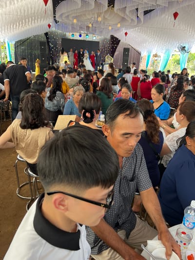 A medium-shot, slightly tilted photograph captures a lively indoor gathering, likely a wedding reception, at Bình An, Vietnam. Numerous guests, primarily adults of various ages, are seated at tables under a elaborately decorated ceiling. The venue features white, ruffled fabric draping the ceiling, interspersed with string lights and hanging red lanterns, creating a festive atmosphere. Natural light enters from an open side, indicating it is daytime, though artificial lighting also illuminates the space.

In the foreground, a man with dark hair and a patterned dark blue shirt is seen looking towards the left, with a younger person wearing glasses partially visible in the bottom right, looking in the same direction. Guests in the midground and background are engaged in conversation or eating. One cropped view confirms the event as a wedding, showing a bride in a white gown and a groom on a stage at the far end. The stage includes a large, decorated archway and what appears to be a multi-tiered wedding cake.

Tables hold water bottles, with one clearly displaying the text "Nước Uống Tinh Khiết Number One," along with plates, glasses, and cutlery. Clothing ranges from casual to more formal attire. Other visible objects include large fans for air circulation, metal chairs, and speakers positioned near the stage. The overall scene depicts a joyous and well-attended celebration.