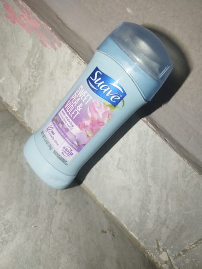 This is an image of a Suave Sweet Pea & Violet anti-staining invisible solid antiperspirant deodorant. The deodorant stick is light blue with a clear cap and features a label with floral imagery. The text on the label is mostly purple and white and includes "SWEET PEA & VIOLET," "anti-staining invisible solid," "antiperspirant deodorant," "Cruelty-Free," and "48HR protection." The bottom of the label also indicates "NET WT 2.6 OZ (74g)."

The deodorant stick is lying on a textured surface that appears to be concrete or stone, possibly a floor or a step. There are some reddish-pink marks on the surface near the top left of the image. The lighting suggests it may be indoors or in an area with artificial light, as there are distinct shadows. No people or specific activities are visible. The overall impression is that of a product photograph.