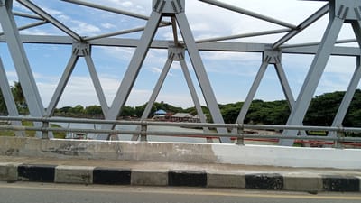 The image shows a view through the metal trusses of a bridge in Neuboknaleung, Indonesia. The bridge's gray metal structure dominates the foreground, with diagonal and horizontal beams forming a geometric pattern against a blue sky with scattered white clouds. Below the bridge, a body of water is visible, with several boats docked or moored near a waterfront area. Along the bank, there are buildings with colorful roofs, trees, and what appear to be parked motorcycles. The water is calm, reflecting the sky and surrounding vegetation. The edge of the bridge railing and the concrete barrier with black and white markings are also visible in the lower part of the image, suggesting the perspective is from a road or walkway on the bridge. The overall scene depicts a peaceful, somewhat rural waterfront environment with a functional bridge as a central element.