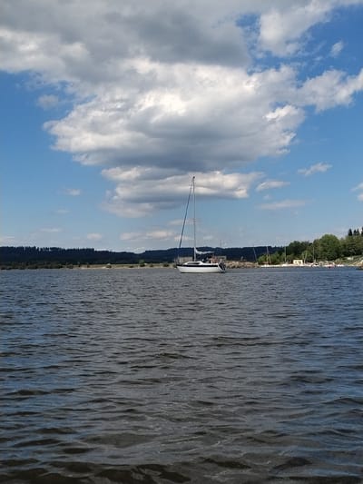 A wide-angle, eye-level, outdoor shot shows a lake with a sailboat in the foreground. The sailboat is white with a dark hull and a tall mast with sails furled. The sailboat is positioned in the center of the frame and appears to be docked or anchored. In the background, a dense forest lines the shore of the lake. The sky is blue with scattered white clouds. The water is dark and rippled, suggesting a slight breeze. The image has a peaceful and serene atmosphere. The specific location of Radslav, Czechia is not identifiable from the image itself.