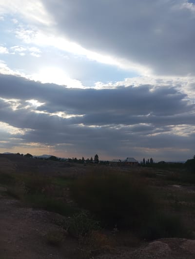 The image depicts an overcast sky with sunbeams breaking through dark clouds, illuminating a rural landscape. In the distance, low hills are visible, and a few trees and buildings, including what appear to be houses under construction, are scattered across the terrain. The foreground consists of rough, dry ground with sparse vegetation. The scene suggests a rural setting, possibly on the outskirts of the city of Kızıl-Bel, Kyrgyzstan, given the architectural style and the topography. There are no people or animals visible in the frame.