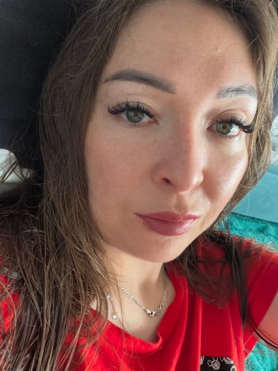 A close-up selfie shows a woman with long, brown, wet hair, a black hat, and a red t-shirt. She has green eyes, dramatic false eyelashes, and wears light pink lipstick. A silver necklace with a heart-shaped pendant rests on her collarbone. The background includes a teal-colored surface, possibly a couch or chair. The image appears to have been taken indoors, and no specific location in Старые Туймазы, Russia, can be identified.