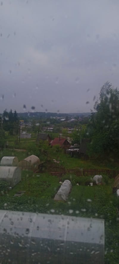 This image depicts a rural scene in Kudymkar, Russia, on a cloudy, overcast day, as evidenced by the raindrops on the window through which the photograph was taken. The foreground is dominated by a large, elongated greenhouse structure made of translucent plastic and metal framing, partially obscured by water droplets. In the middle ground, several smaller, arched greenhouses are visible, covered in white material, suggesting a garden or agricultural setting. Beyond the greenhouses, a cluster of traditional wooden houses with red roofs is nestled among lush green trees and vegetation. A rustic wooden fence runs through the yard, separating different areas. In the distance, a village or townscape with more buildings and trees can be seen under the grey sky, stretching towards a dense forest line. The overall atmosphere is one of a quiet, perhaps slightly melancholic, rural day, marked by the rain. There are no people or visible text in the image. The time of day is difficult to determine precisely due to the overcast conditions, but it appears to be daytime.