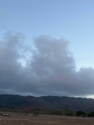 This image depicts a landscape scene featuring rolling hills or mountains in the background, under a sky with scattered clouds. The foreground consists of dry, barren land with sparse vegetation and some scattered debris. The overall lighting suggests either early morning or late evening. The location is identified as San Antonio, Venezuela.