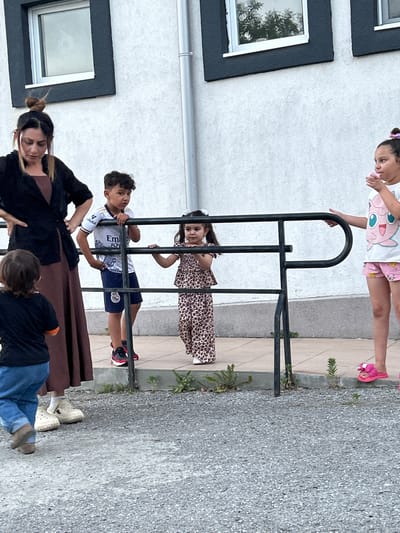 This image shows a group of people, primarily children, interacting near a black metal railing. In the background, a white building with dark-framed windows is visible. The ground is a mix of gravel and a light-colored paved surface.
At the top of the frame, a young girl wearing a white t-shirt with a pink cartoon character and pink shorts with white polka dots is holding onto the railing. She has her hair tied up with a pink accessory and is wearing pink slides. Below her, another young girl in a leopard print dress is also holding onto the railing, looking towards the camera.
In the middle section, a boy in a white t-shirt with blue and white accents, and dark shorts, is also holding onto the railing. His t-shirt has visible text that appears to be "Emirates Fly Better" and "LA LIGA". He is wearing dark athletic shoes with red and black accents.
At the bottom of the frame, an adult woman with her hair in a bun is wearing a dark jacket over a pink top and a brown skirt or dress. She is looking down and appears to be holding something. Next to her, another person, possibly a child, is wearing a black t-shirt and blue jeans, with their legs visible. The overall scene suggests a casual outdoor gathering. The lighting indicates it is daytime, and the weather appears to be pleasant.