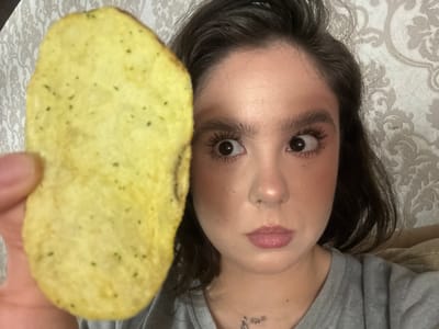 In a close-up, medium shot, a young woman with dark hair is holding a large potato chip in her left hand, close to the camera, obscuring part of her face. Her eyes are wide and looking directly at the camera, with a slightly surprised or humorous expression. She is wearing makeup, including mascara and blush. She is wearing a grey t-shirt and a silver necklace. The background consists of a patterned wallpaper. The image does not contain any information to suggest the location is in Raivka, Ukraine.
