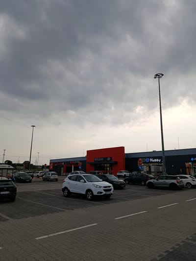 The image depicts a parking lot outside a commercial building, likely a shopping center, with the city of Łagów, Poland, as its location. The sky is overcast with heavy grey clouds. In the foreground, a paved parking lot is occupied by several cars, including a black Citroen, a white Hyundai, and a silver Mini Cooper. In the middle ground, the building features a large orange section with the word "Witamy" (Welcome) and a Carrefour logo, adjacent to a dark grey section with logos for "Reserved," "Hobby," and other businesses. A tall lamppost stands prominently in the mid-right of the image. In the background, a few more cars are visible, and to the far left, a sign for "Carrefour" is present. Electrical pylons and a distant building can be seen on the horizon. The scene appears to be during daylight hours, possibly late afternoon or early evening, given the dim lighting conditions. There are no discernible people in the image performing any specific actions.