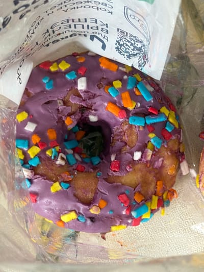 A close-up, top-down shot shows a purple frosted donut with colorful sprinkles. The donut is in a clear plastic bag, and part of a white paper wrapper with text and a QR code is visible in the upper left corner. The donut has a hole in the center, and the frosting is unevenly distributed. Sprinkles of red, yellow, blue, and orange are scattered across the top of the donut. The background is blurred, suggesting it is an indoor setting, possibly a bakery or food establishment. The image does not provide any specific details about the city of Crokhovo, Russia, nor does it depict any people, actions, or interactions. The scene is focused solely on the donut and its immediate packaging.