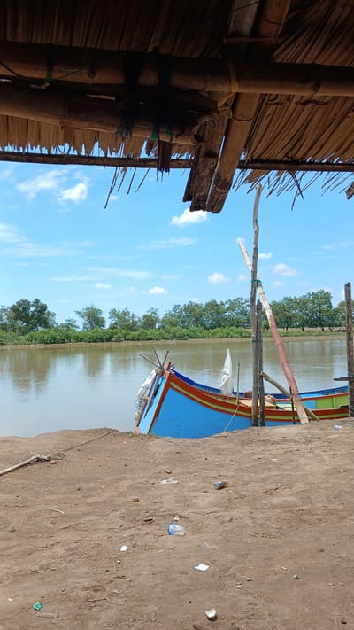 The image depicts a scene along the riverbank in Kualacangkoi, Indonesia. A blue and red fishing boat is moored on sandy ground, its bow pointing towards the viewer. The boat has a white flag attached to its mast and several buoys visible. Behind the boat, a calm, murky river stretches out, lined by dense green foliage and trees on the opposite bank. The sky above is a bright blue, dotted with scattered white clouds. In the foreground, the sandy ground is littered with various pieces of trash, including plastic bottles and wrappers, and a discarded can. Overhanging the scene is the rustic roof of a structure, constructed from bamboo and palm leaves, supported by wooden beams. The overall impression is one of a working river environment, with elements of both nature and human activity, including the presence of pollution.