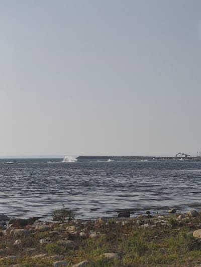 The image depicts a serene coastal scene with a calm body of water in the foreground. The water is a deep blue, with gentle ripples creating a textured surface. Along the shoreline, there are scattered rocks and sparse vegetation, suggesting a natural or undeveloped area. In the distance, a long breakwater extends across the water, punctuated by what appear to be industrial structures, including a crane. Waves are visible crashing against the breakwater, creating white spray. The sky above is a pale, overcast blue, contributing to the overall sense of tranquility. The scene captures a moment of coastal activity near a likely industrial or port facility in the vicinity of Saiyn Şapağatov, Kazakhstan.