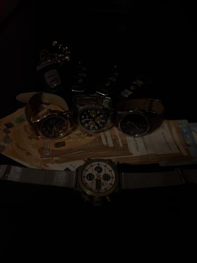 The image displays a dark, top-down still-life arrangement of luxury and personal items on a black surface, likely indoors in Dolni Tsibar, Bulgaria. The scene is lit dramatically, with most details emerging from deep shadow.

The primary subjects are four wristwatches, several Euro banknotes, and multiple car keys or fobs.

The watches vary in style:
1.  One features a brown leather strap and a black dial with "LOTU" branding.
2.  Another is a silver-toned watch with a black dial and chronograph subdials.
3.  A rose gold-toned watch has a brown leather strap, a black dial with Roman numerals, and chronograph functions, bearing "WOVE" branding.
4.  The most prominent watch is silver-toned with a mesh strap, a light-colored dial, chronograph subdials, and a digital display. Visible text on this watch includes "QUARTZ", "DATE", "WATER RESISTANT", "ALARM", "START/STOP", "RESET", and "LIGNT" (possibly part of "LIGHT").

Several Euro banknotes, appearing to be 50 Euro notes, are fanned out beneath the watches. Visible text and serial numbers on these notes include "EC3811301648", "69031504875", "6627943862", "340", and the year "2017". A signature or stylized text "you jande" is also visible on one note.

Multiple car keys and fobs are positioned to the left, largely obscured by shadow, with one key resembling a BMW design. The composition evokes themes of wealth, personal belongings, and time.