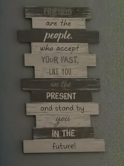 The image shows a piece of wall decor made of wooden planks stacked vertically. Each plank has text printed on it. The text reads: "FRIENDS are the people who accept YOUR PAST, LIKE YOU in the PRESENT and stand by you IN THE future!" The wall decor is mounted on a textured gray wall. The lighting is dim, and the wall decor is slightly angled, with the left side appearing closer to the viewer than the right. There are no people or animals visible in the image. The environment appears to be indoors, likely a home or a business setting. The specific location in the city of Rheinfelden (Baden), Germany, cannot be determined from the visual information provided in the image.
