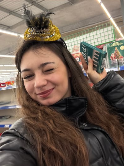 An image, rotated counter-clockwise, depicts a young woman in what appears to be a retail store aisle in Lampertheim, France. She has long, wavy dark brown hair and is wearing a black faux leather jacket. A festive golden, sequined mini-hat or headband with black feathers adorns her head, suggesting a celebratory atmosphere. Her eyes are closed as she smiles broadly, indicating joy or amusement.

She holds up a small, rectangular turquoise-blue box in her right hand. The text on the box is clearly visible, reading: "nail it!", "24 NAILS WITH GLUE", "SEA KELP", "POINTED SHAPE", and "CLASSIC DESIGN", indicating the product is artificial nails.

The background features shelves stocked with various products, illuminated by artificial lighting typical of an indoor store environment. A green metal grid, possibly part of a display or shopping cart, is visible behind her. Among the items on the shelves, a box featuring a Christmas tree design is discernible, with text identifying it as a "Christmas Scenery Nightlight" by "LX". Another partially visible box displays the text "DIAM". The woman's cheerful expression and festive accessory suggest a playful moment while shopping.