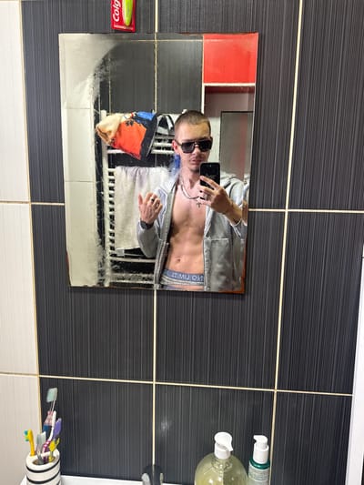 The image depicts a bathroom setting. A man with sunglasses is reflected in a mirror, appearing to take a photo of himself. He is wearing an unzipped silver jacket, revealing his torso and the waistband of his underwear. In the reflection, a patterned towel hangs behind him. The bathroom features a mix of black and white tiled walls, with visible toiletries such as soap dispensers and toothbrushes. There is no indication that this image was taken in Vetren Dol, Bulgaria.