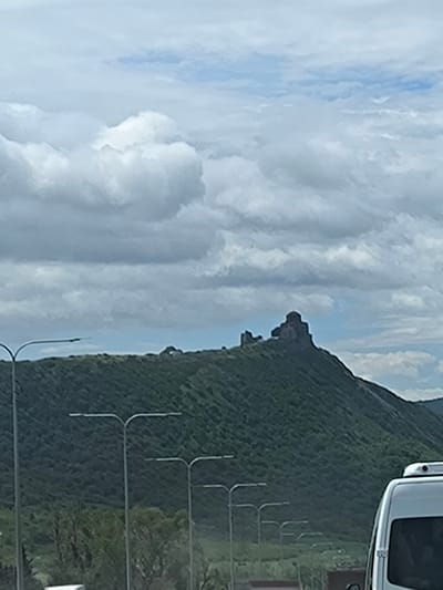 The image shows the Jvari Monastery situated atop a green, tree-covered mountain near the city of Tsitsamuri, Georgia. The sky is filled with grey and white clouds. In the foreground are roadside street lamps and a portion of a white vehicle.