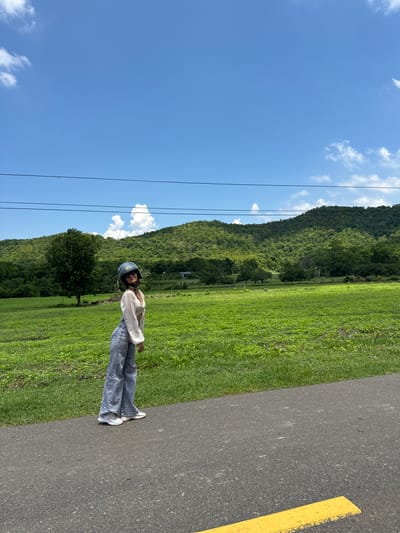 A young woman stands on an asphalt road in Ham Thuan Bac, Vietnam, facing slightly away from the camera and looking over her shoulder. She is wearing a white crop top, blue flared jeans, and a blue helmet. The road has a yellow painted line on its right side. Behind the road is a vibrant green field, followed by a dense forest covering a rolling hillside, and a bright blue sky with scattered white clouds. Three parallel black wires stretch horizontally across the upper portion of the image.